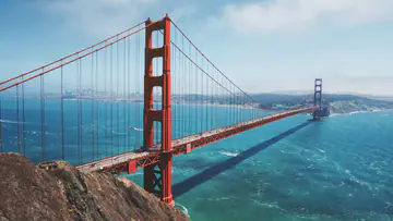 Orange suspension bridge over a bay, seen from a cliff
